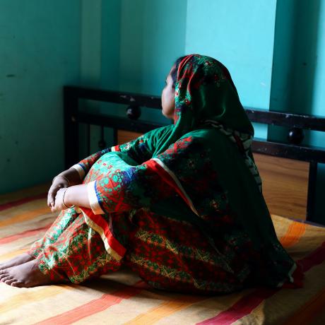 A woman sitting on a bed faces away from the camera, with her hands around her knees. 